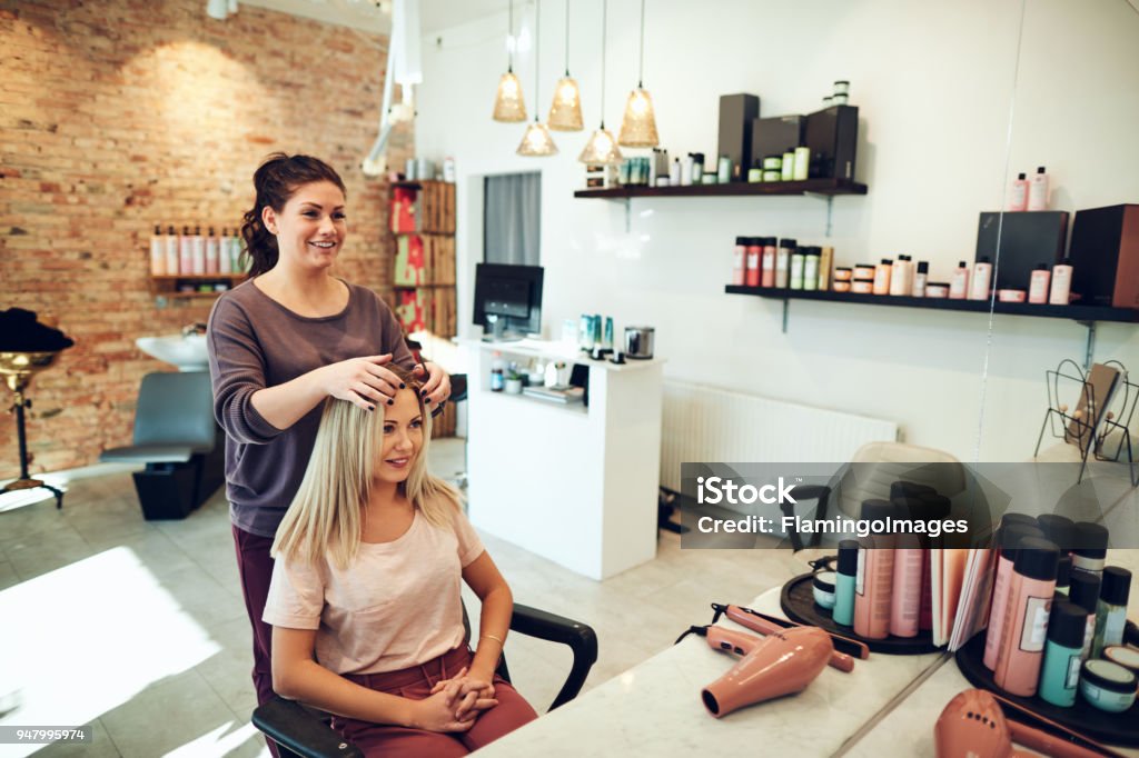 Young woman talking with her hairstylist during a salon appointment Young blonde woman smiling and looking at her reflection in a mirror while sitting in a salon chair during an appointment with her hairstylist Hair Salon Stock Photo