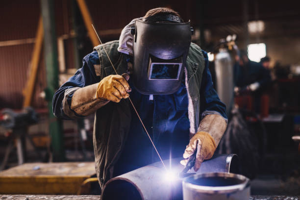 istock Welder in protective uniform and mask welding metal pipe on the industrial table while sparks flying. 947996020