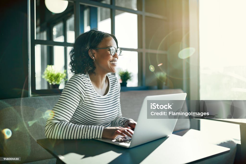 Smiling young African woman working online with her laptop Smiling young African woman wearing glasses looking out of a window while sitting at a table working online with a laptop Office Stock Photo