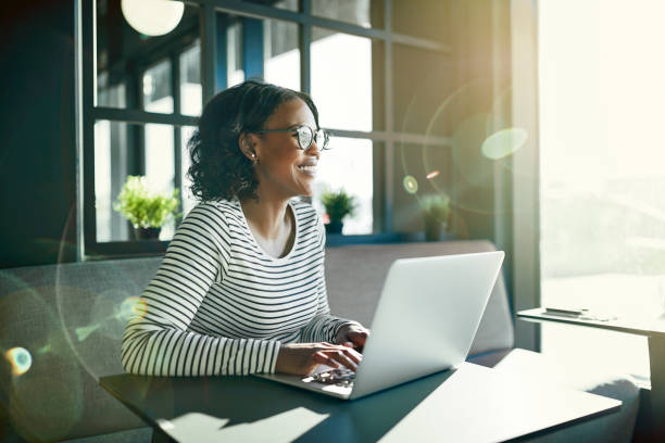 Smiling young African woman working online with her laptop Smiling young African woman wearing glasses looking out of a window while sitting at a table working online with a laptop sunny window stock pictures, royalty-free photos & images
