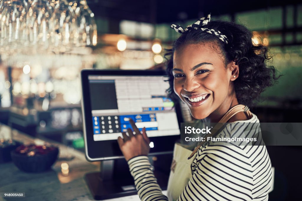 Smiling African waitress using a restaurant point of sale terminal Smiling young African waitress wearing an apron using a touchscreen point of sale terminal while working in a trendy restaurant Point Of Sale Stock Photo