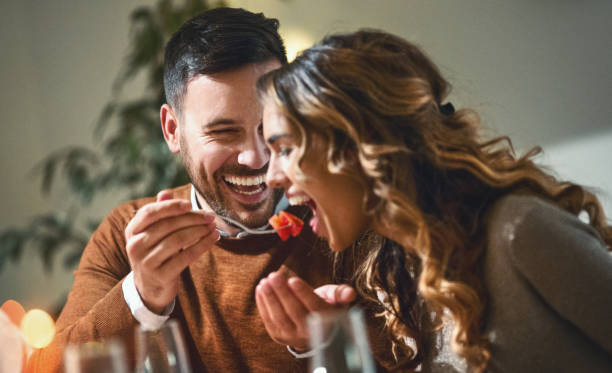 Dinner party. Closeup of mid 20's couple having fun during dinner party. The guy is feeding his girls with some chopped fruit, both laughing. romance stock pictures, royalty-free photos & images