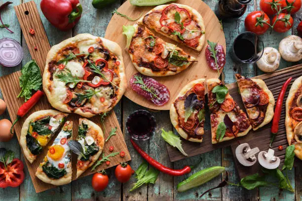 Flatlay of abundant pizza party. Rustic homemade pizzas with salami, eggs and vegetables served with red wine and raw vegetables on shabby wooden background overhead view