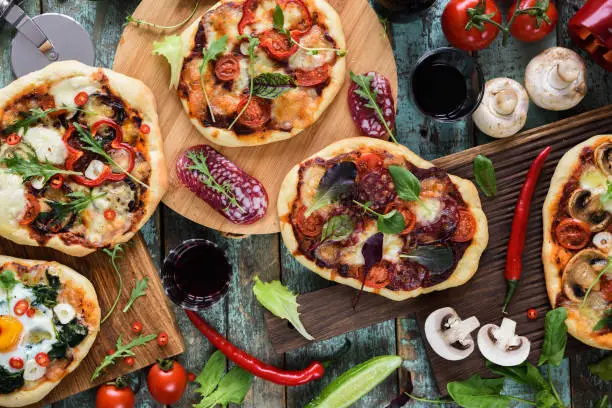 Pizza and wine party. Homemade rustic pizzas and raw ingredients served with red wine on oak chopping boards over shabby blue background top view