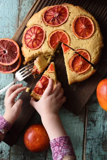 Baby taking piece of homemade cornmeal cake with blood oranges on dark oak board over shabby rustic blue background overhead view