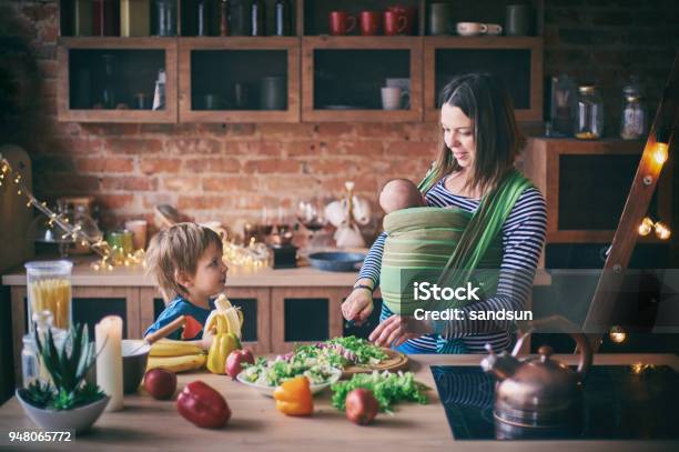 Photo libre de droit de Heureux Jeune Famille Belle Mère Avec Deux Enfants Adorable Garçon Préscolaire Et Bébé En Écharpe Cuisine Ensemble Dans Une Cuisine Ensoleillée banque d'images et plus d'images libres de droit de Mère