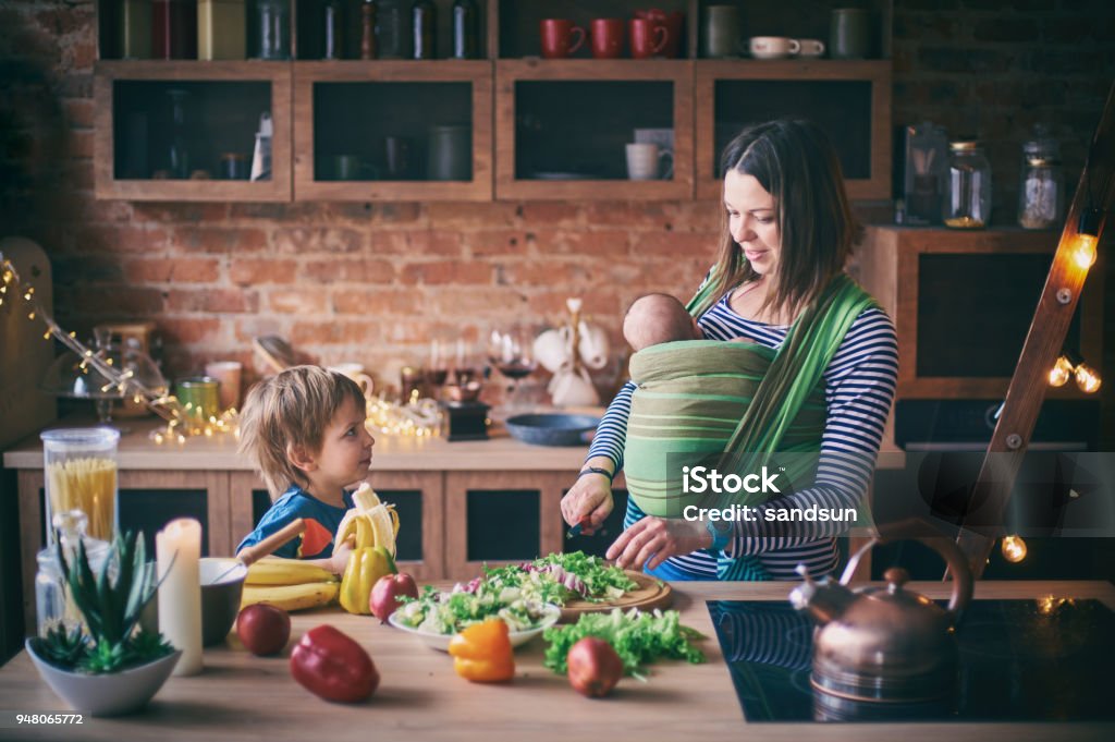 Happy young family, beautiful mother with two children, adorable preschool boy and baby in sling cooking together in a sunny kitchen. Happy young family, beautiful mother with two children, adorable preschool boy and baby in sling cooking together in a sunny kitchen. Vintage style. Mother Stock Photo