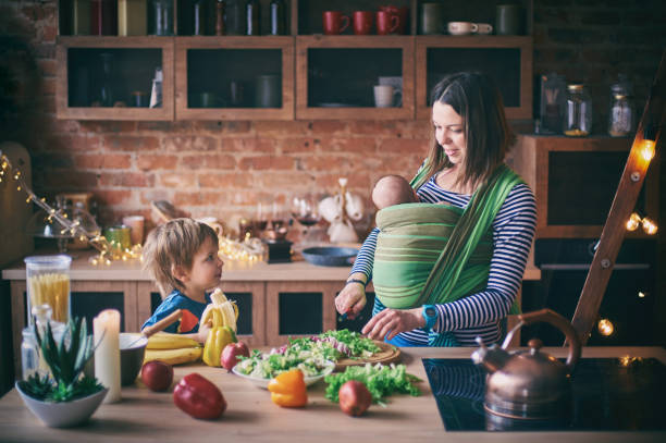 happy young family, beautiful mother with two children, adorable preschool boy and baby in sling cooking together in a sunny kitchen. - breakfast family child healthy eating imagens e fotografias de stock