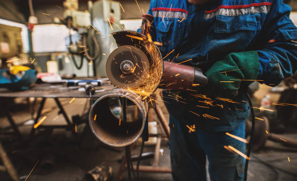 istock Fabric worker in protective uniform cutting metal pipe on the work table with an electric grinder in the industrial workshop. 948106292