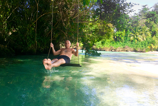 Smiling senior man sitting on a swing over a tropical river under a tree in Jamaica