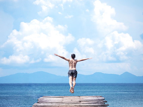 Back view of asian man jumping off wooden bridge into blue sea background. Flying freely to the sky. Summer vacation lifestyle.