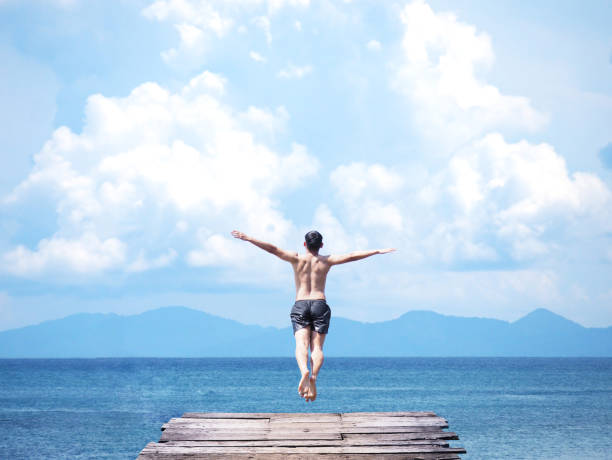 asian man jumping off wooden bridge into blue sea Back view of asian man jumping off wooden bridge into blue sea background. Flying freely to the sky. Summer vacation lifestyle. people jumping sea beach stock pictures, royalty-free photos & images