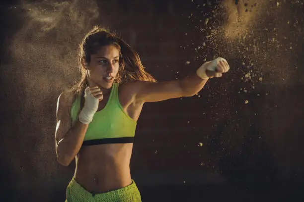 Concentrated athletic woman doing a boxing punch on a training. Dust is around her.