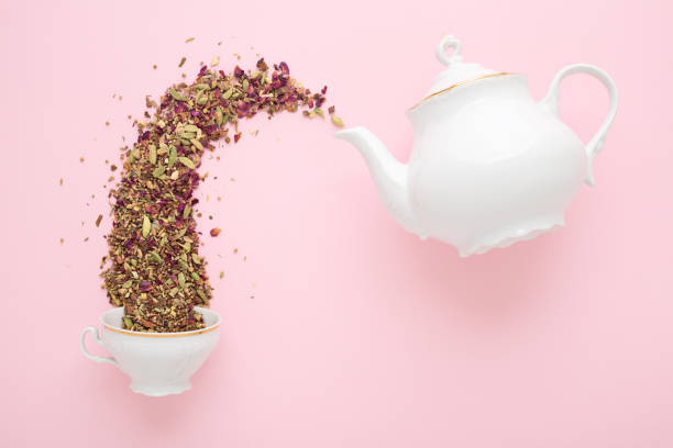Dry tea pouring from teapot into cup on pink Dry herbal tea pouring from white porcelain teapot into cup on pink surface. Flat lay. Tea time concept. pink tea stock pictures, royalty-free photos & images