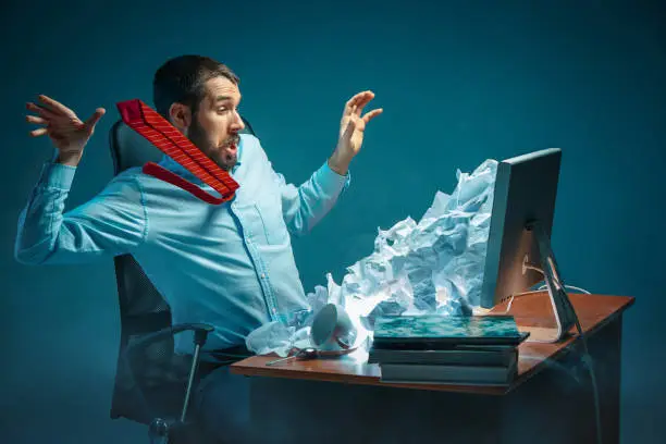 Young stressed handsome businessman working at desk in modern office shouting at laptop screen and being angry about e-mail spam. Collage with a mountain of crumpled paper. Business, internet concept