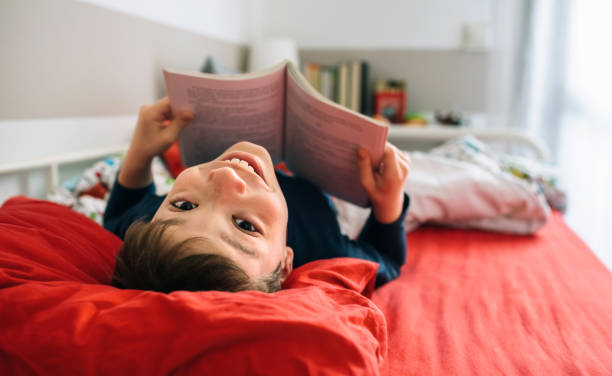 macchina fotografica dall'aspetto ragazzo mentre legge un libro - bed child smiling people foto e immagini stock