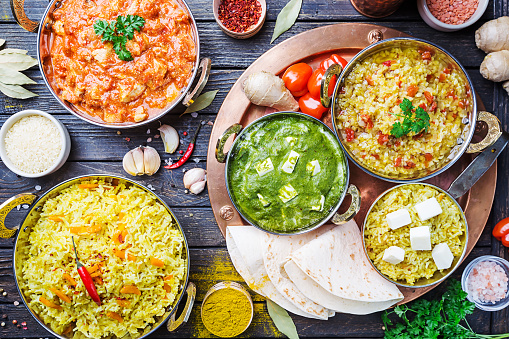 Different bowls with assorted indian food on dark wooden background, top view. Dishes and appetizers of indian cuisine. Chicken, curry rice, lentils, paneer, chapati and spices.