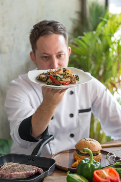 istock Professional chef in uniform holding spaghetti plate in his hand with food on table foreground. 948498378