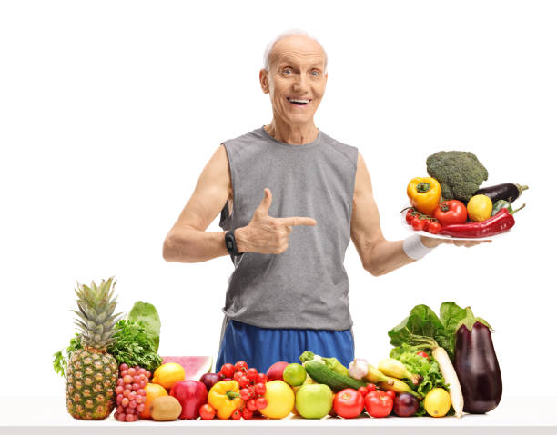 istock Senior behind a table with fruit and vegetables holding a plate and pointing 948510706