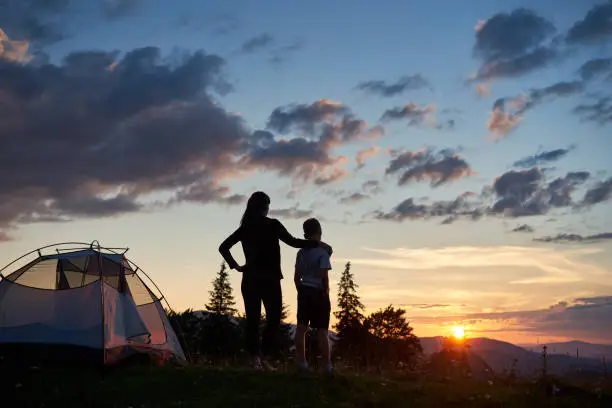 Photo of Rear view of mother hugs her son near tent on top of mountain enjoying view of sun
