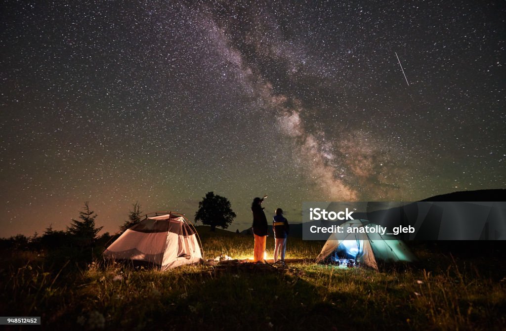 Mother and son tourists resting at camping in mountains at night sky full of stars and Milky way Back view mother and son tourists resting at camping in mountains, standing beside campfire and two tents, looking at night sky full of stars and Milky way, enjoying night scene. Woman pointing at sky Camping Stock Photo