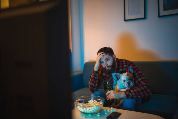 istock Young bearded men watching sport on television with his dog 948556550