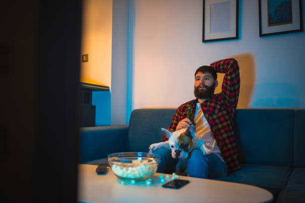 istock Young bearded men watching sport on television with his dog 948556582