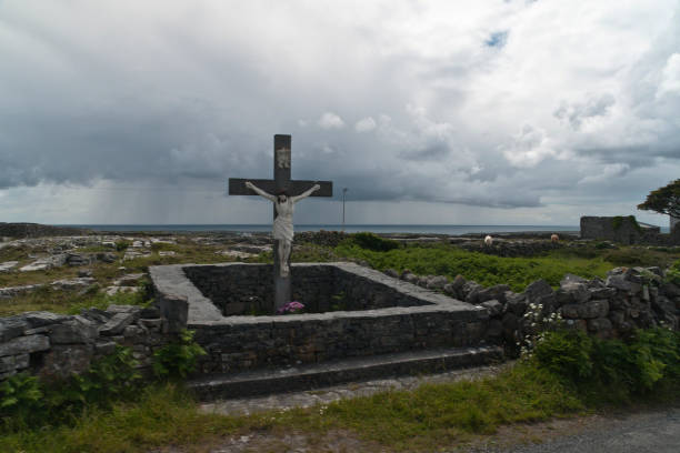 Roadside Shrine and Island Landscape, Inishmore, Aran Islands, County Galway, Ireland A roadside shrine on Cottage Road, Inishmore.  A large crucifix set with dry stone walls with cut flowers.  The walls are the native limestone.  It is a spring (early June) afternoon and there are fern and wildflowers.  The white flowers are Greater Burnet saxifrage (Scientific Name: Pimpinella major).  In the distance are more dry stone walls around fields, a stone shed, feeding horses and the sea, being Galway Bay, storm clouds with distant rain.  Aran Islands, County Galway, Ireland. michael stephen wills aran stock pictures, royalty-free photos & images