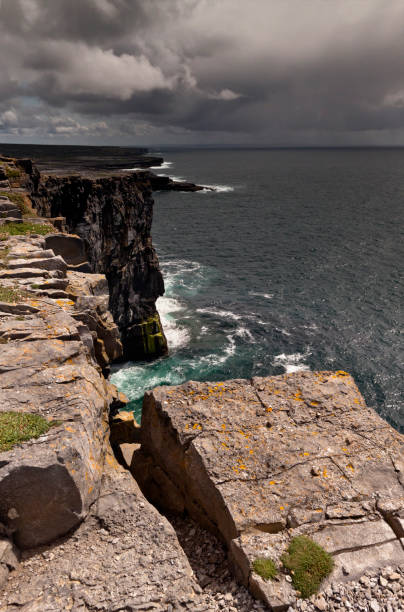 View east, southeast from the Cliff Edge of the Ancient fort wall of Dun Aonghasa (Dun Aengus), Inishmore, Aran Islands, County Galway, Ireland Breaking waves below the dramatic cliffs of the ancient fort Dun Aonghasa (Dun Aengus), in the distance the karst landscape of Inishmore with clouds of an approaching storm over Galway Bay.  Aran Islands, County Galway, Ireland. michael stephen wills aran stock pictures, royalty-free photos & images