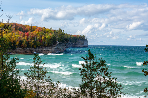 An Upper Peninsula forest creates an autumn background at Chapel Beach in northern Michigan. Lake Superior waves crash on the beach and white puffy clouds dot the blue sky.