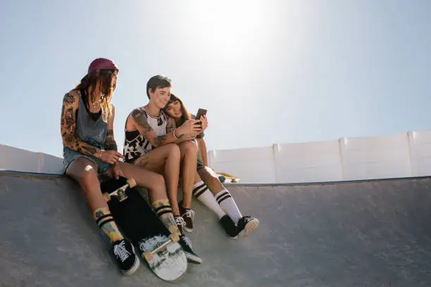 Group of female friends sitting on ramp at skate park and looking at mobile phone. Three women skaters using smart phone together at skate park.