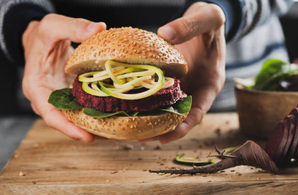 young man with a beet burger sandwich closeup of a young caucasian man with a beet burger sandwich in his hands, next to a rustic wooden table and a sliced beet veggie burger stock pictures, royalty-free photos & images