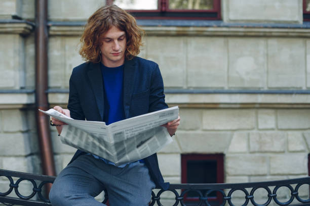 istock Young reddish man reading newspaper near old style building 948675482