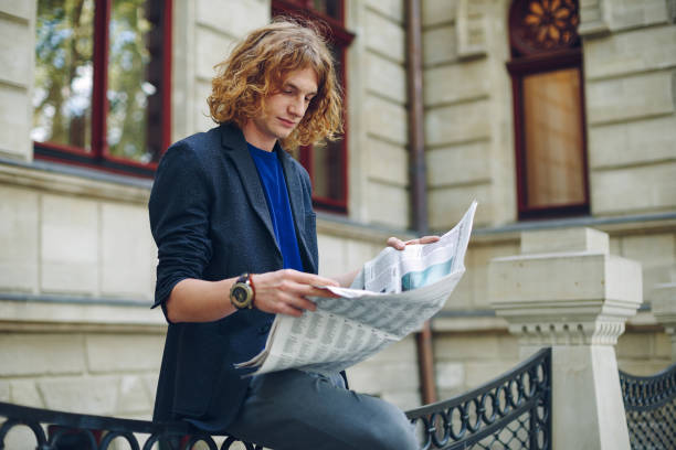 istock Young reddish man reading newspaper near old style building 948675502