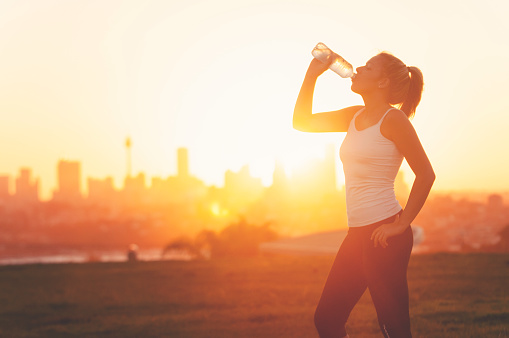 Silhouette of a woman drinking form a cold water bottle. She is exercising at sunset or sunrise. City of Sydney in the background. Copy space.