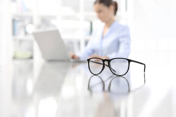 eyeglasses leaning on desk and woman working on computer at office in background eyeglasses leaning on desk and woman working on computer at office in background lens optical instrument stock pictures, royalty-free photos & images