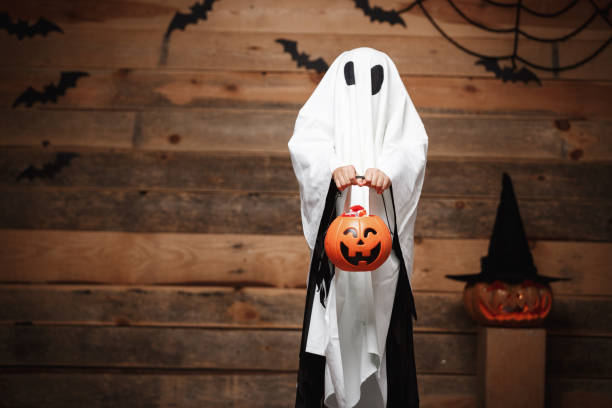 istock Halloween Concept - little white ghost with halloween pumpkin candy jar doing trick or treat with curved pumpkins over bats and spider web on Wooden studio background. 948729112