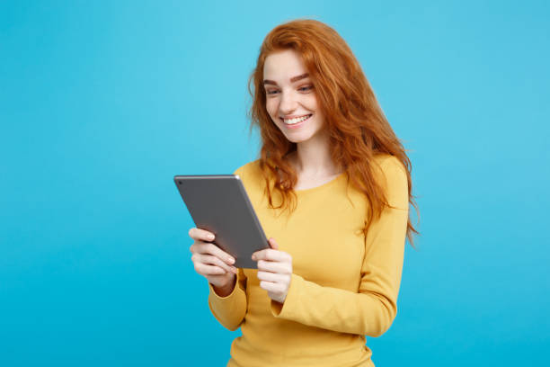 istock People and Technology Concept - Close up Portrait young beautiful attractive tender ginger redhair girl happy smiling on digital table with wining something. Blue Pastel Background. Copy space. 948732832