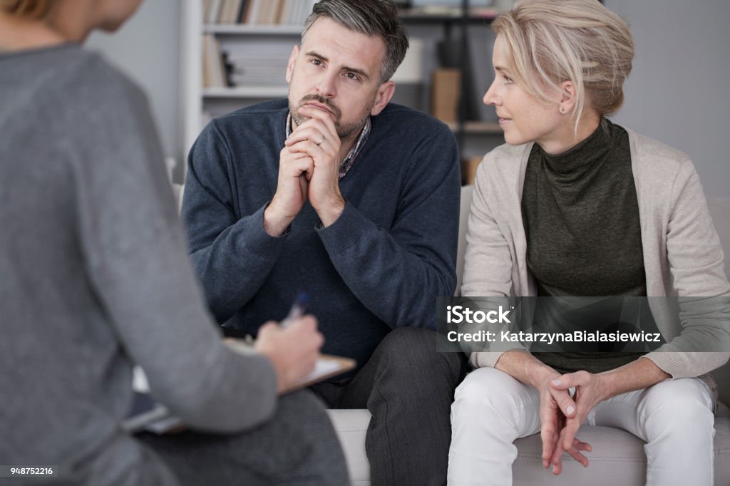 Wife supporting husband in therapy Wife supporting her husband in therapy with the man listening curious to counsellor in gray office Couple - Relationship Stock Photo