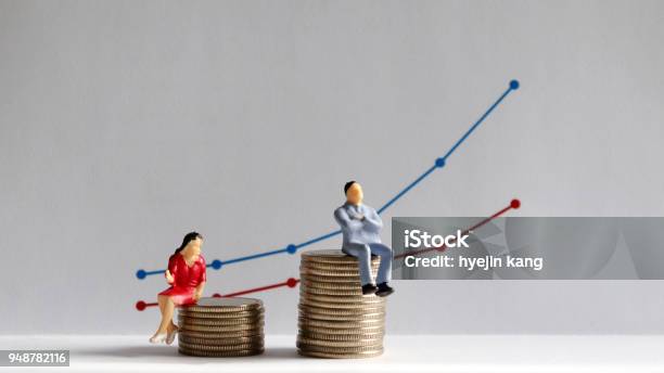 The Concept Of Gender Pay Gap A Miniature Man And A Miniature Woman Sitting On Top Of A Pile Of Coins At Different Heights In Front Of A Bar Graph Stock Photo - Download Image Now