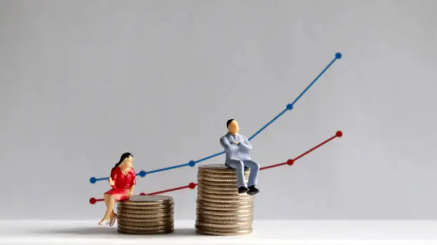 Photo of The concept of gender pay gap. A miniature man and a miniature woman sitting on top of a pile of coins at different heights in front of a bar graph.