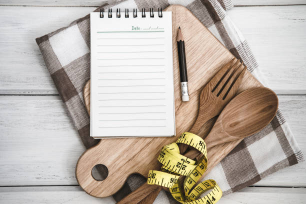 istock Notepad on chopping board with wooden fork and spoon and measuring tape on white table ,a  recipes food or diet plan for healthy habits shot note background concept 948785596