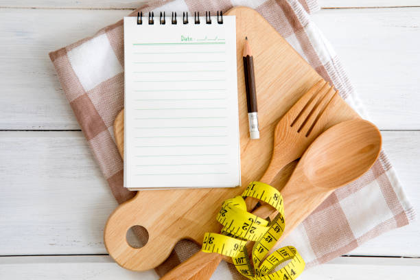istock Notepad on chopping board with a wooden fork and spoon and measuring tape on white table , recipes food or diet plan for healthy habits shot note background concept 948785852