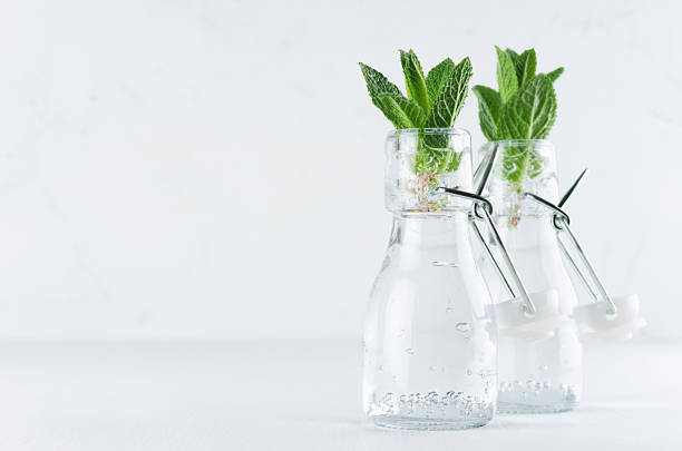 istock Elegant soft white summer background - young mint leaves in transparent bottles with sparkling bubbles on light wood table, copy space. 948867908