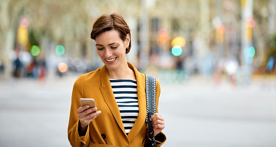 Beautiful mid adult woman texting on smart phone in city. Smiling female pedestrian is wearing yellow long coat. She is walking on street.