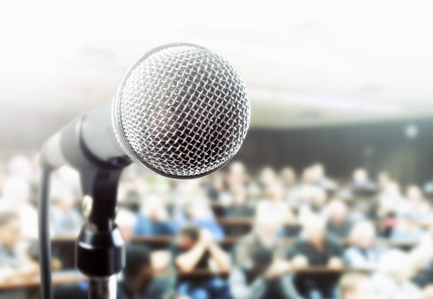 A vocal microphone waits for the speaker in a lecture hall, a large defocused audience in the background.