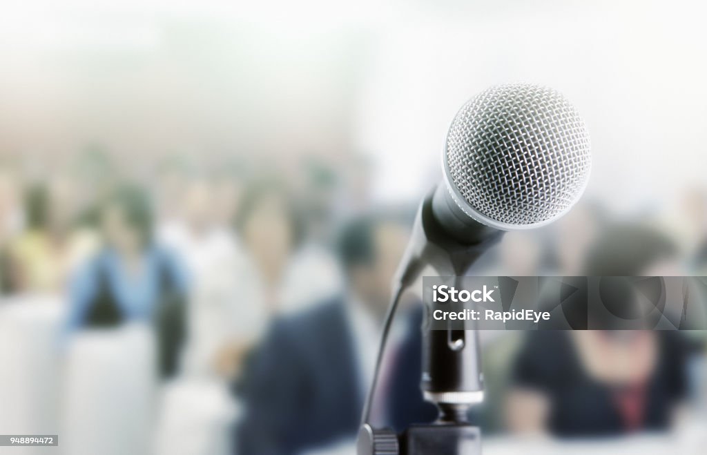 Microphone awaits public speaker at seminar Audience sits waiting for the speaker at a conference. The microphone is in the foreground. Convention Center Stock Photo