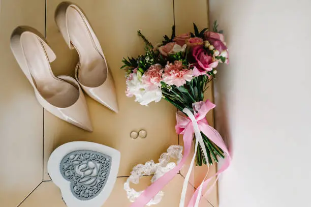 Wedding accessory. Classic lacquered shoes, bouquet bride's of pink flowers roses and greens, two silver rings, garter, box in form of a heart lying on pastel table. Close up. flat lay. top view.