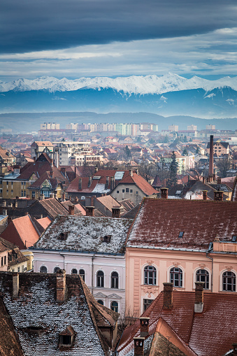 Cityscape scene depicting the ancient medieval city skyline of Sibiu, a city in the Transylvania region of Romania. We can see snow covered roofs of old houses in the foregroud, which give way to the majestic snow-capped Carpathian mountains on the horizon. Sibiu is an ancient medieval city which served as European Capital of Culture in 2007. Room for copy space.