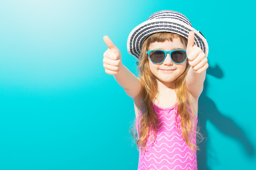 Little happy girl in beach outfit with hat and sunglasses holding thumbs up on blue background.